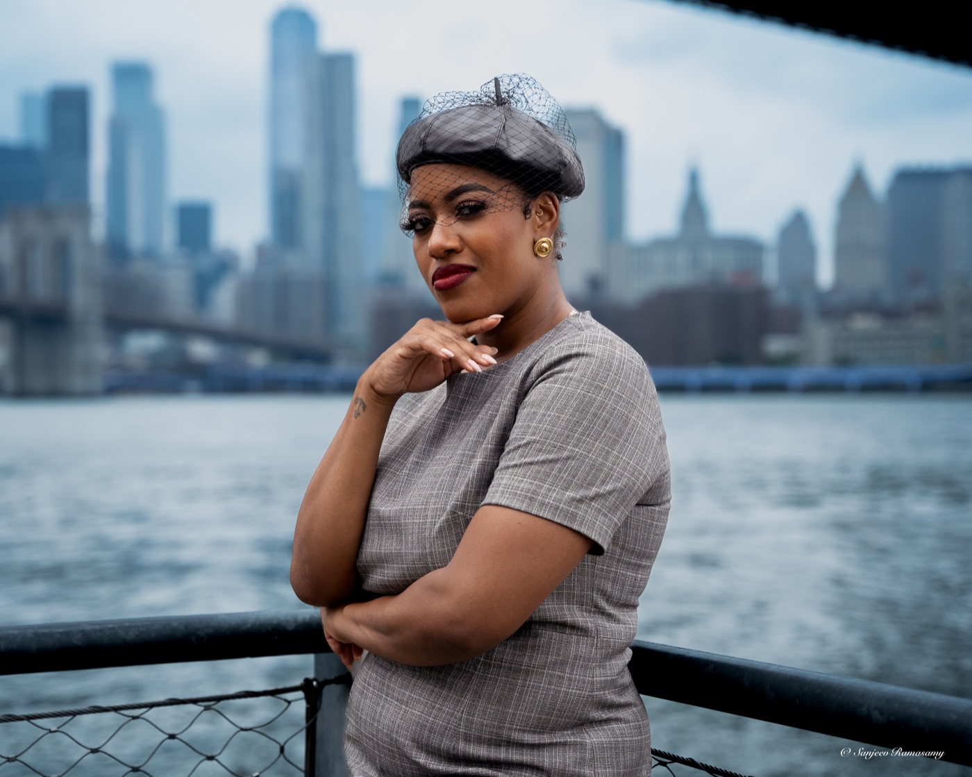 Woman in a grey veiled beret and checked dress leaning on a waterfront railing, a city skyline across the river.