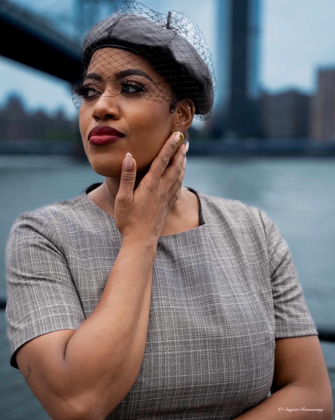 Woman in a grey veiled beret and checked top with a hand to her cheek, gazing away, a bridge and river behind her.