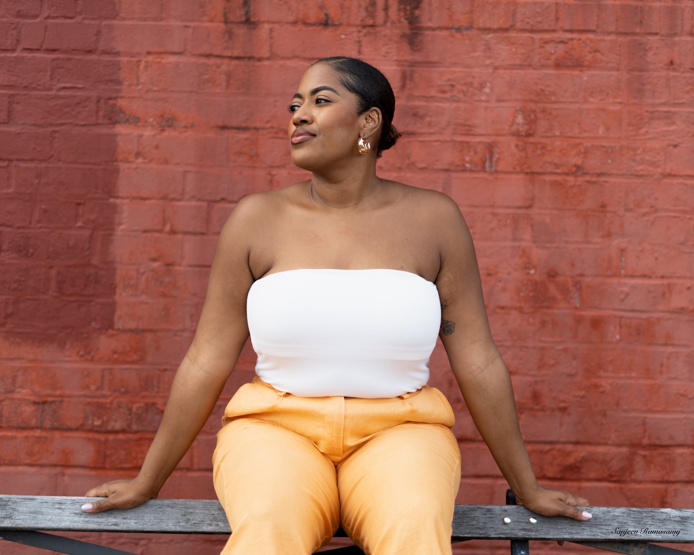 Woman in a white strapless top and peach trousers seated on a bench in front of a red brick wall, looking to the side.