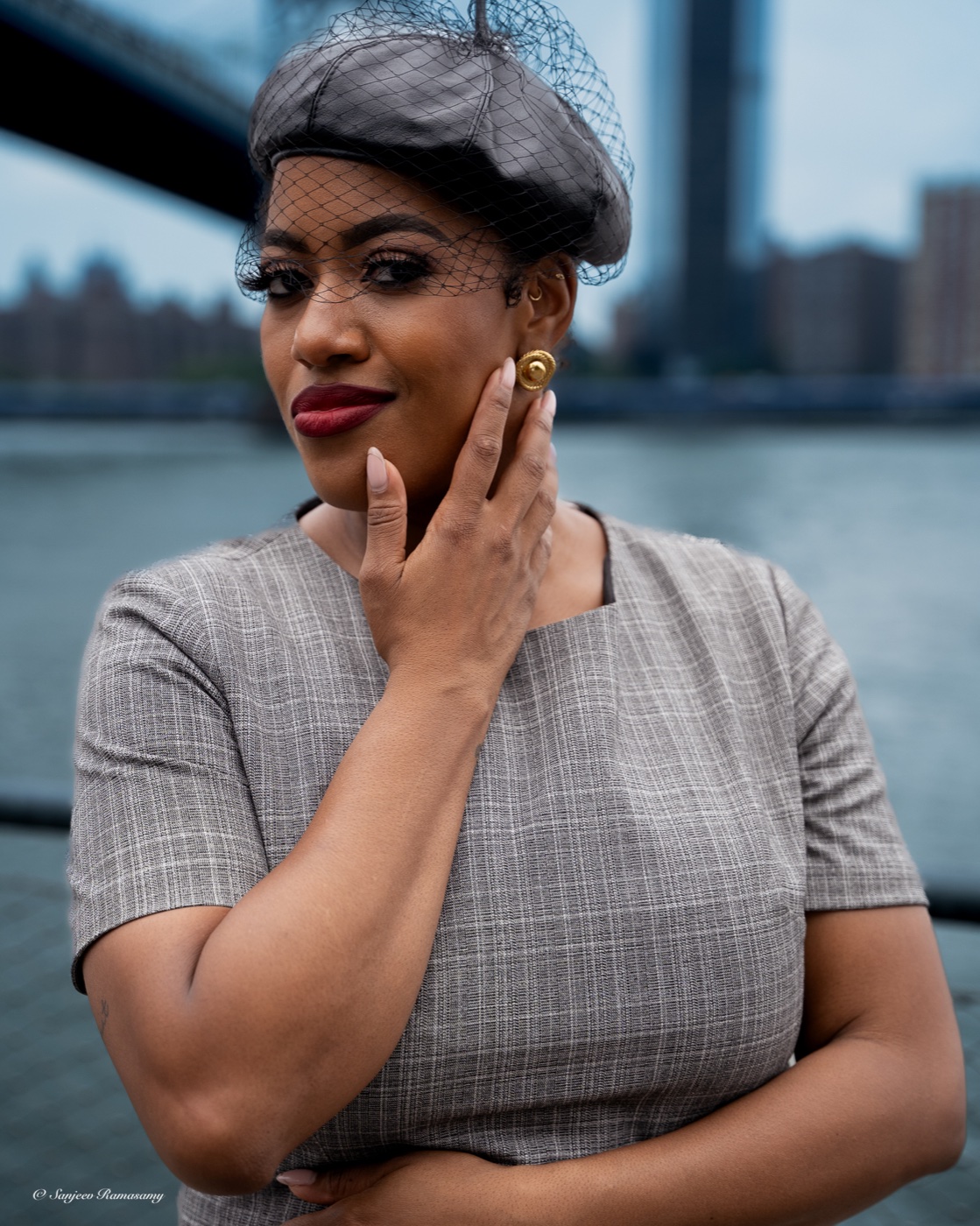 Woman in a grey beret with a small net veil and a grey checked top touching her cheek, with a river and bridge behind her.