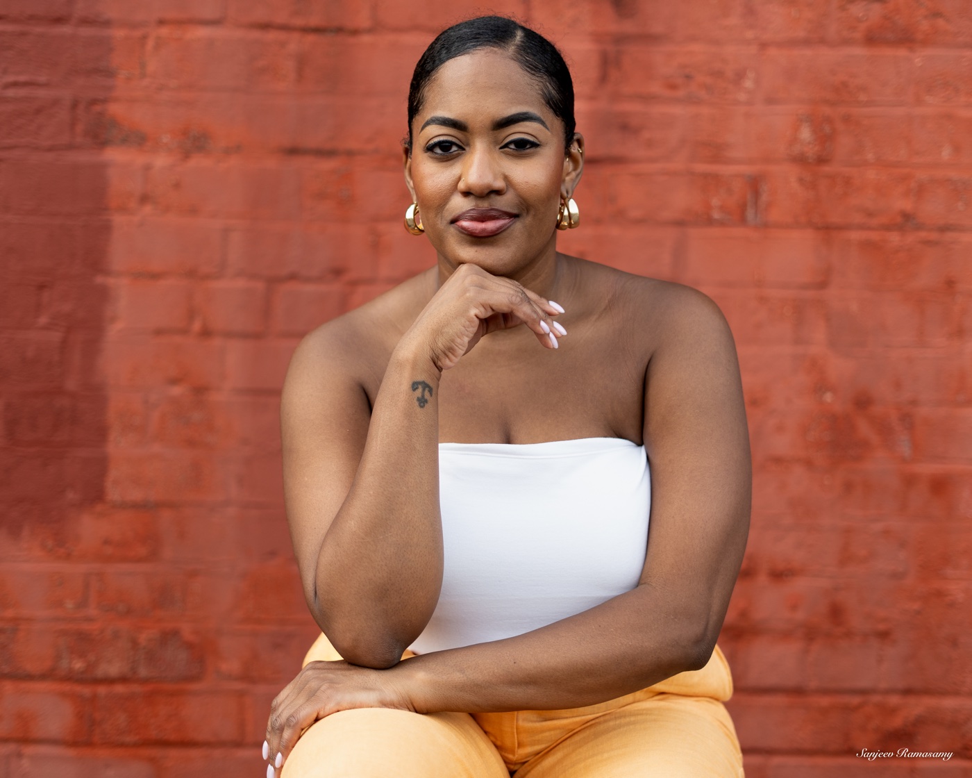Smiling woman in a white strapless top and gold earrings resting her chin on her hand in front of a red brick wall.