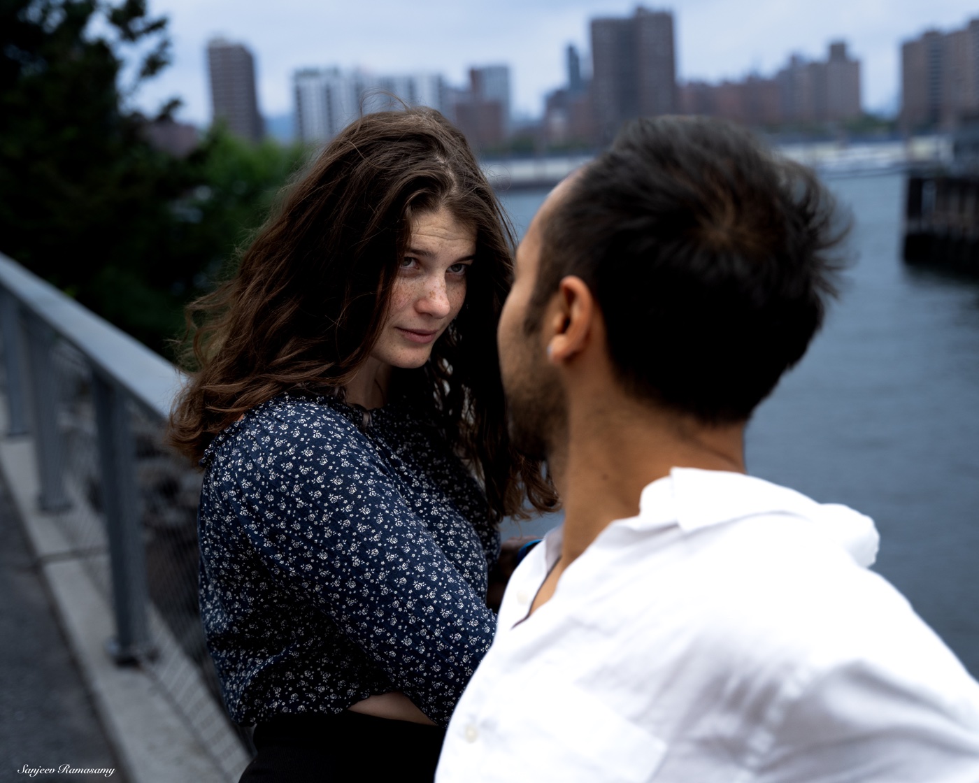 Woman with long wavy hair in a patterned blouse looking toward a man in a white shirt beside a riverside railing, a city skyline behind them.