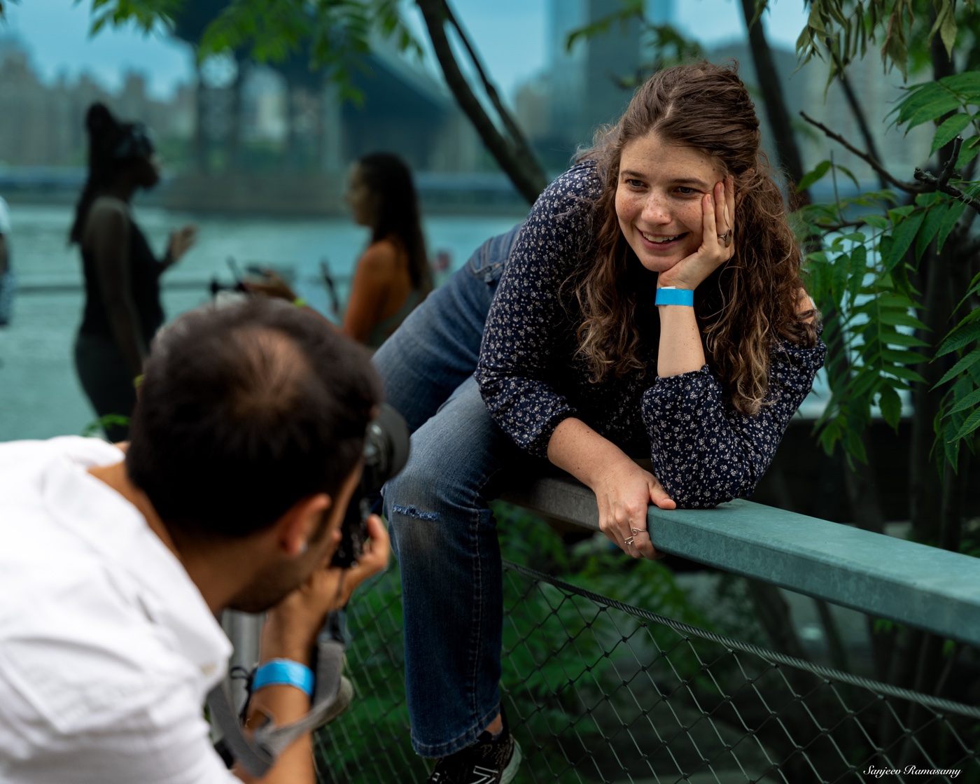 Smiling woman leaning on a railing while a photographer in the foreground photographs her by the water.