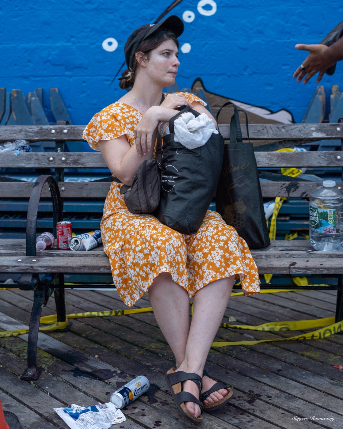 Woman in an orange floral dress and a cap sitting on a wooden bench with bags beside her, in front of a bright blue mural.