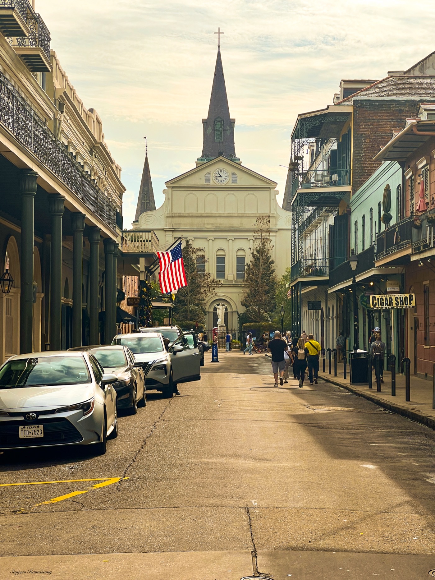 Narrow street lined with balconied buildings and parked cars, leading to a white church with tall spires; an American flag hangs from a balcony.