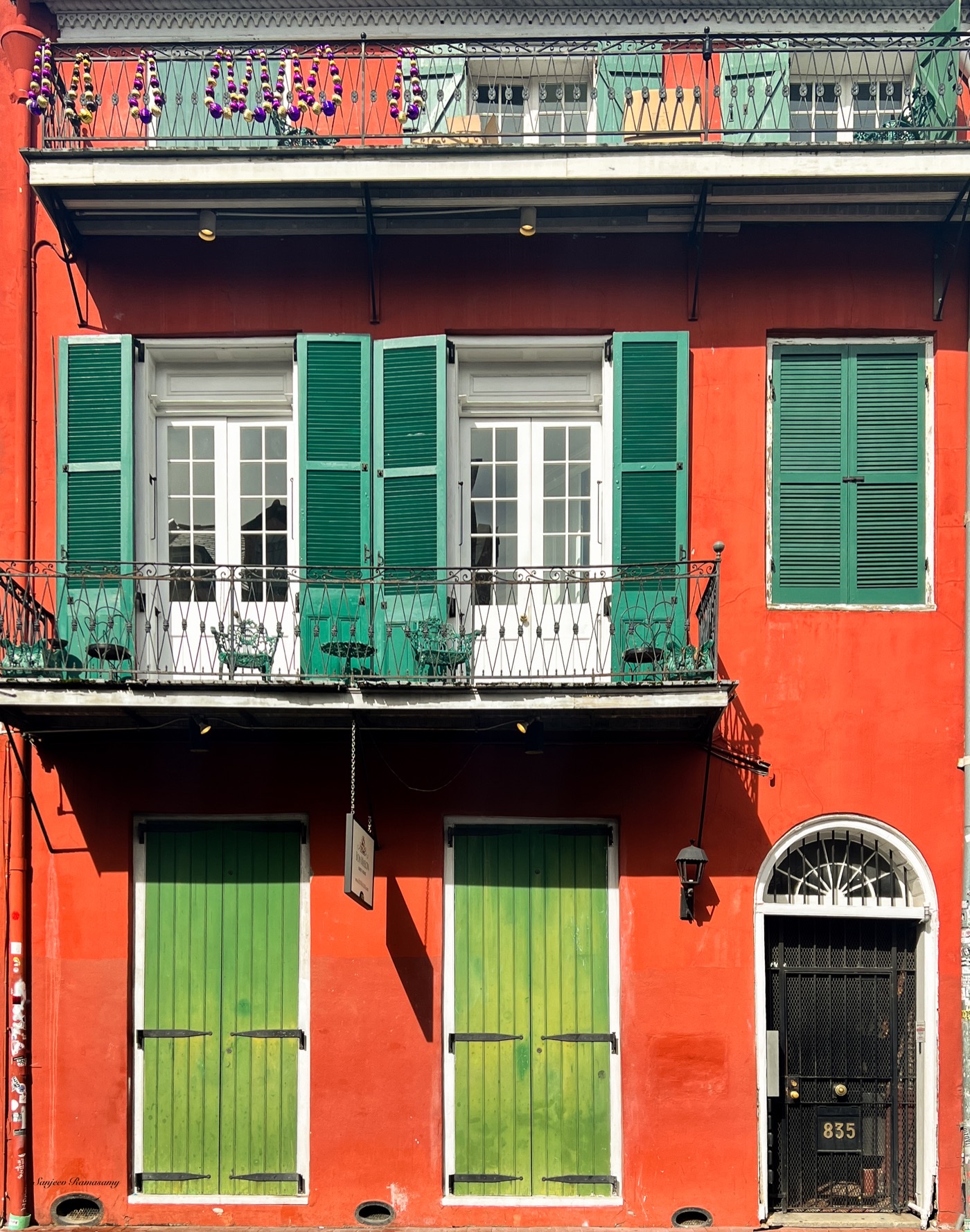 Red townhouse with green shutters, a white balcony railing and two tall green doors at street level.