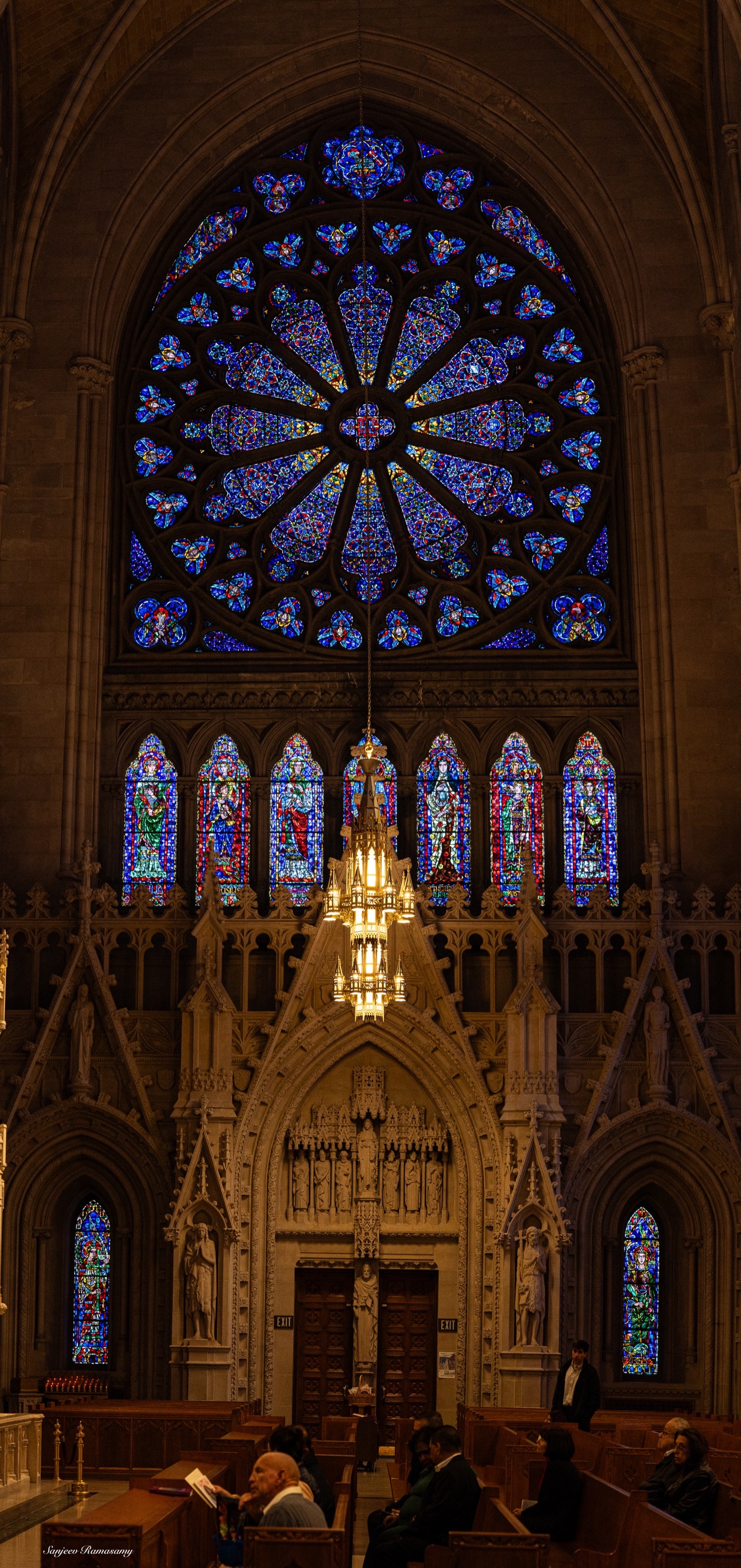 Interior of a Gothic church with a large blue stained-glass rose window above a carved wooden screen, lit by warm lamps.