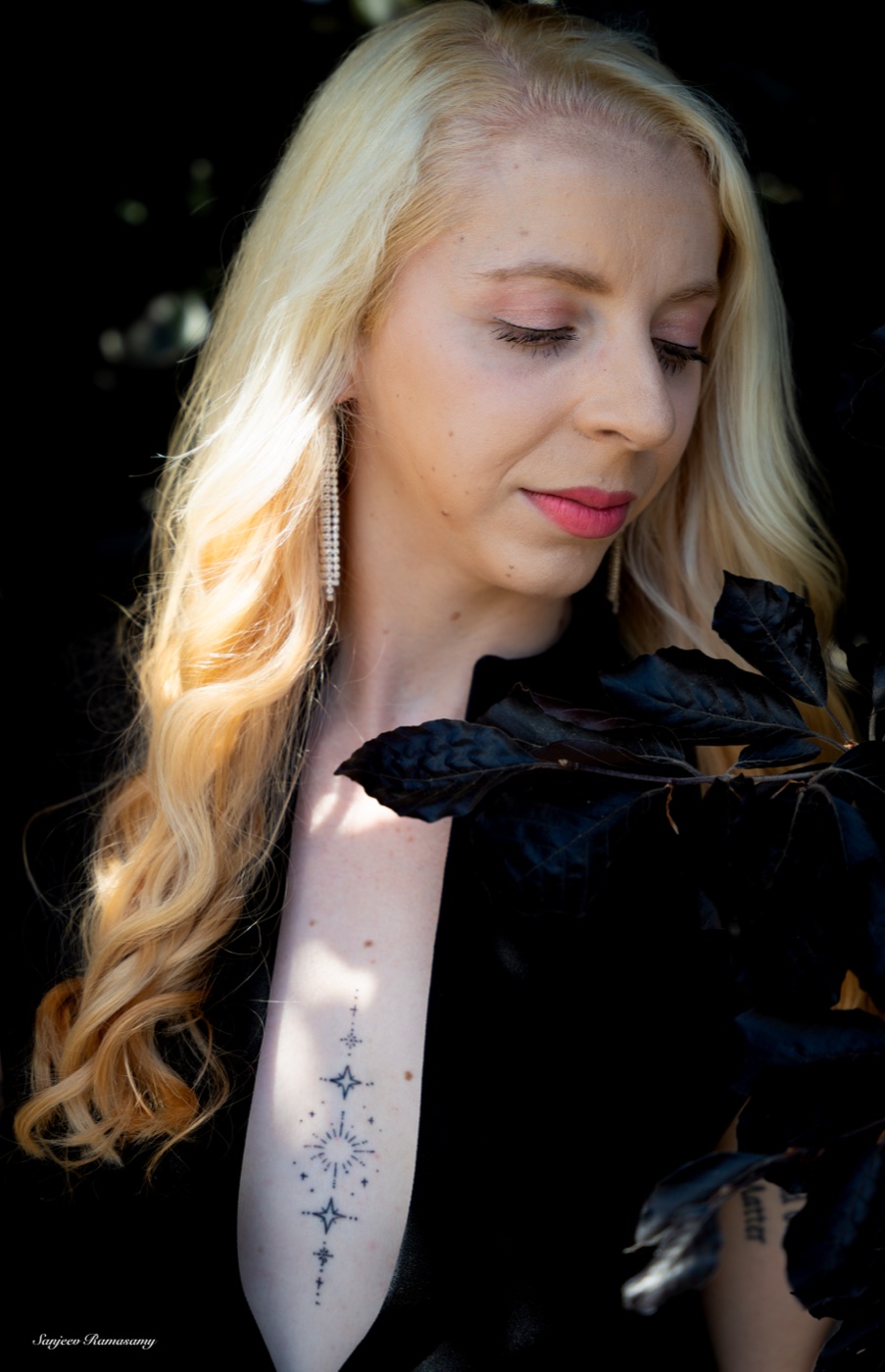 Woman with long wavy blonde hair looking down in warm side light, dark leaves around her.