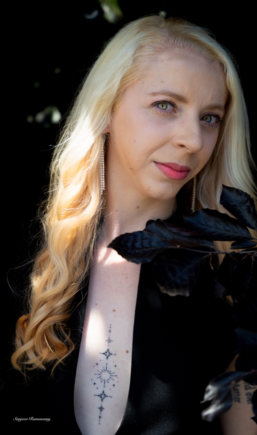 Portrait of a woman with long wavy blonde hair and a fine-line tattoo, looking at the camera in warm light against a dark background.