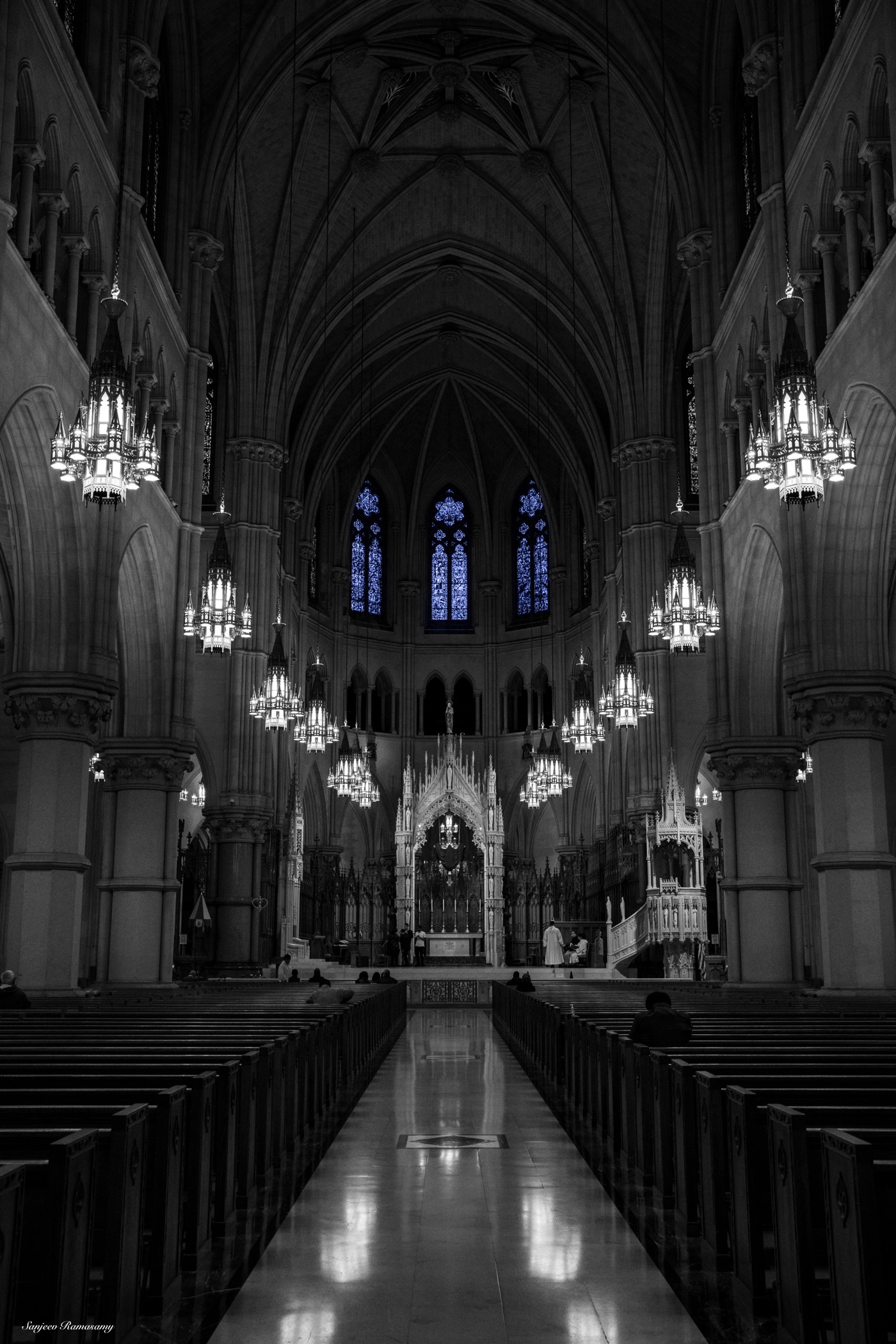 Black-and-white view down the long nave of a Gothic church, with rows of pews, hanging chandeliers and stained-glass windows.