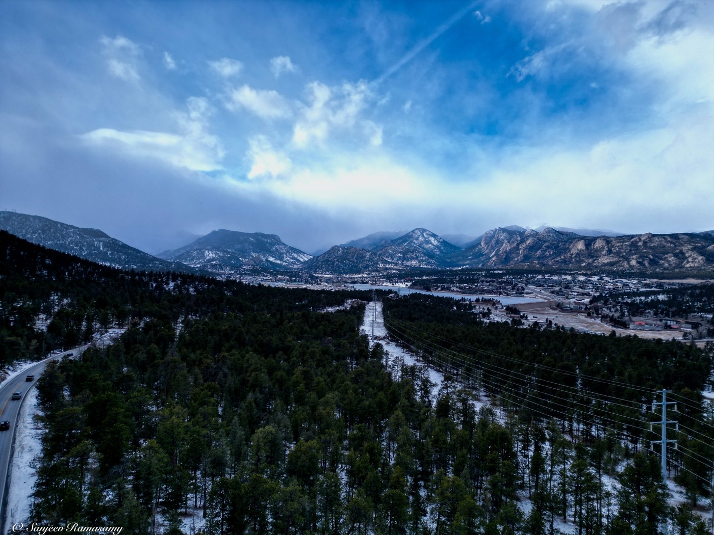 Aerial view over snow-dusted pine forest and a road leading toward distant mountains beneath streaky clouds.