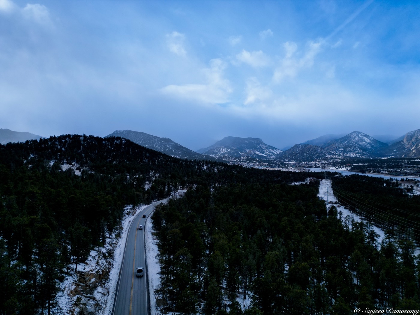 Aerial view of a road cutting through snowy pine forest toward a range of mountains under a bright blue sky.