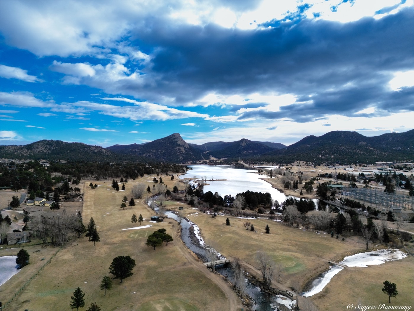 Aerial view of a lake, a winding river and open meadows below snow-streaked mountains under dramatic clouds.