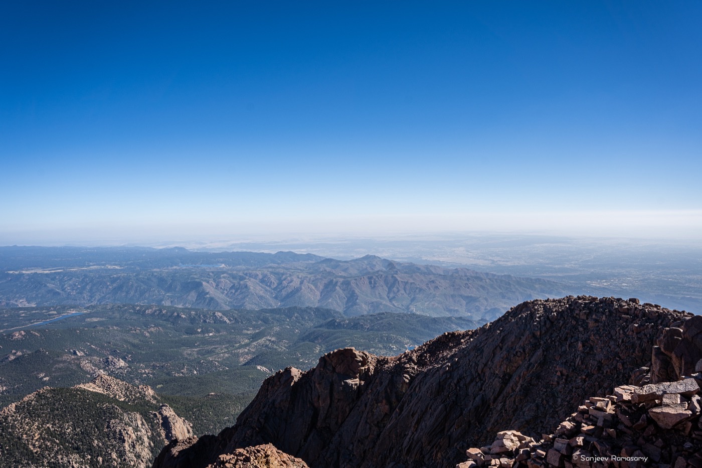 View from a rocky summit over layered mountain ranges fading into a hazy horizon under a clear blue sky.