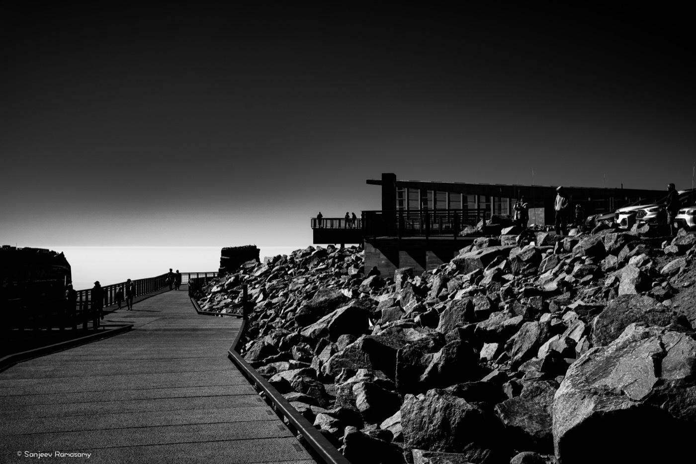 Black-and-white view of a railed walkway and a building on a boulder-strewn summit against a dark sky.