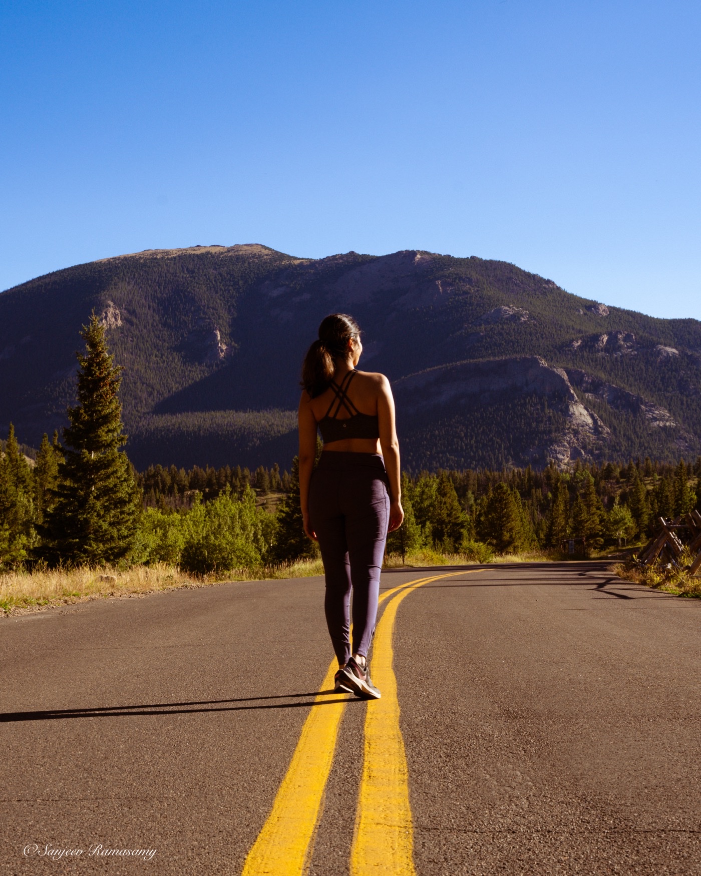 Woman in athletic wear walking away along the yellow centre line of an empty mountain road.
