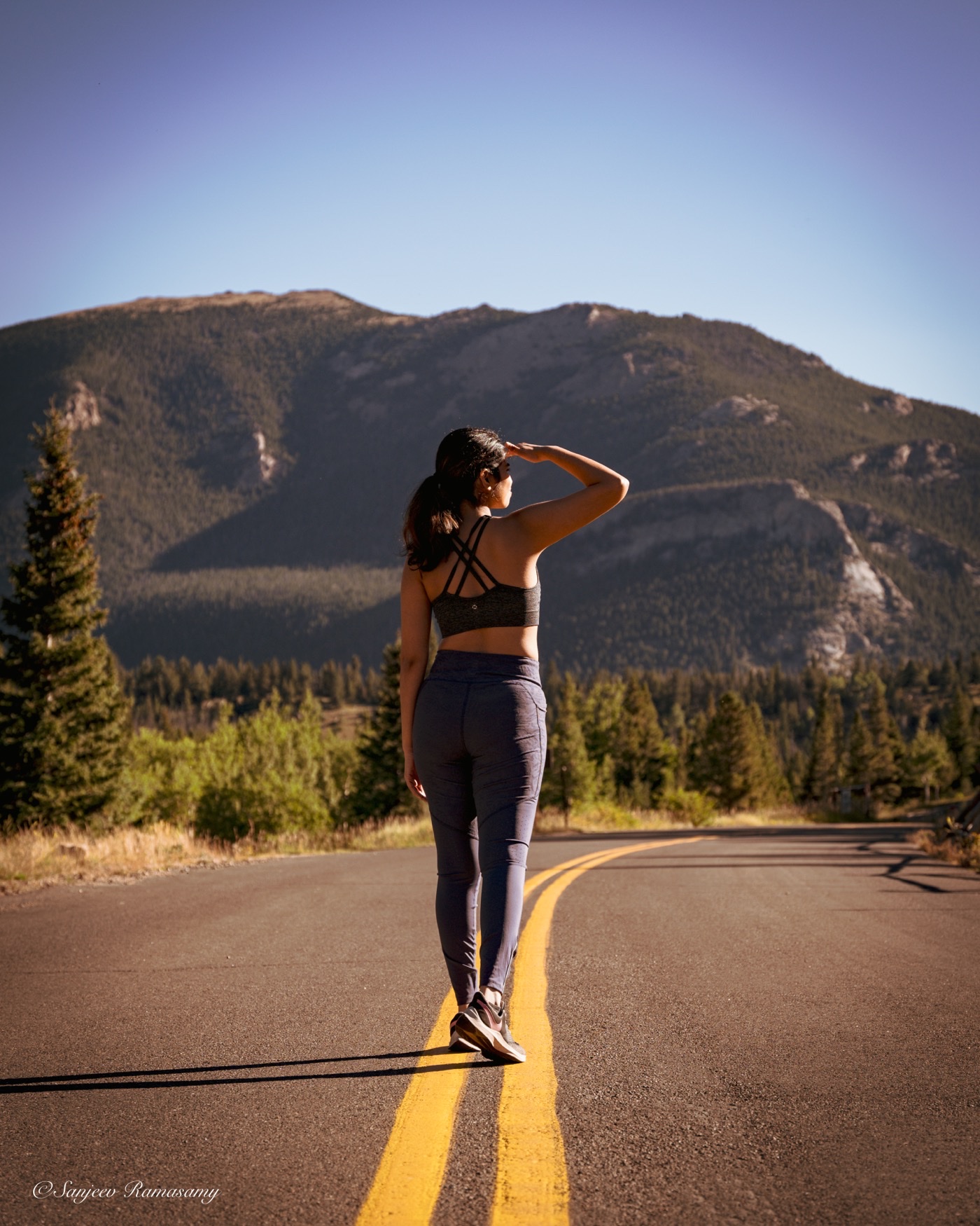 Woman in athletic wear standing on the yellow centre line of an empty road, shading her eyes as she looks toward the mountains.