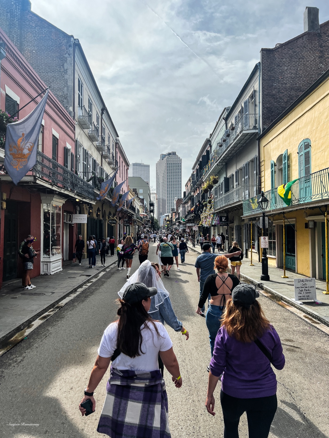 People walking down a street lined with balconied, pastel-coloured buildings, with tall towers in the distance under a cloudy sky.