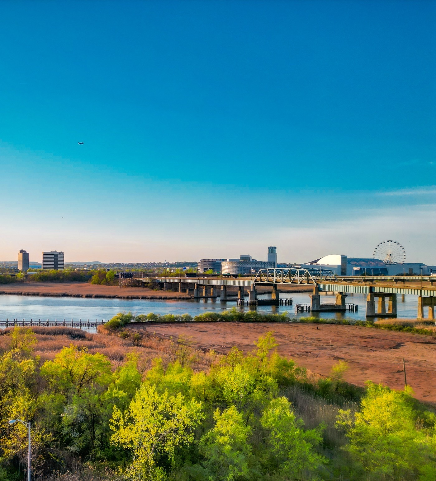 Aerial view over marsh grasses toward a waterfront with a bridge and a Ferris wheel on the horizon.