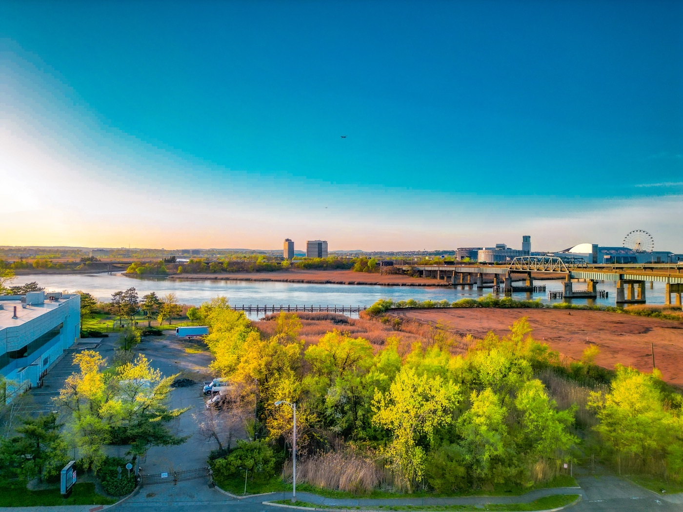Aerial view of marshland and trees along a waterfront, with a bridge, low buildings and a Ferris wheel on the horizon under a clear blue sky.