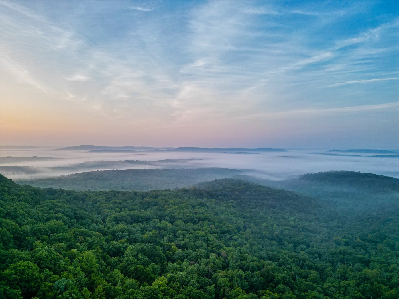 Aerial view of rolling forested hills with low fog drifting through the valleys under a streaked blue sky.