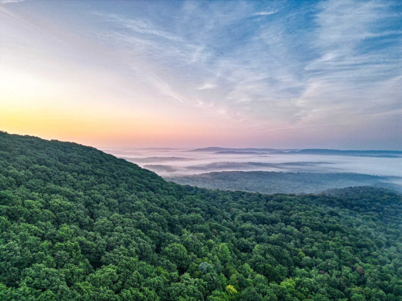 Aerial view over forested hills with fog in the valleys and a pastel sunrise sky.