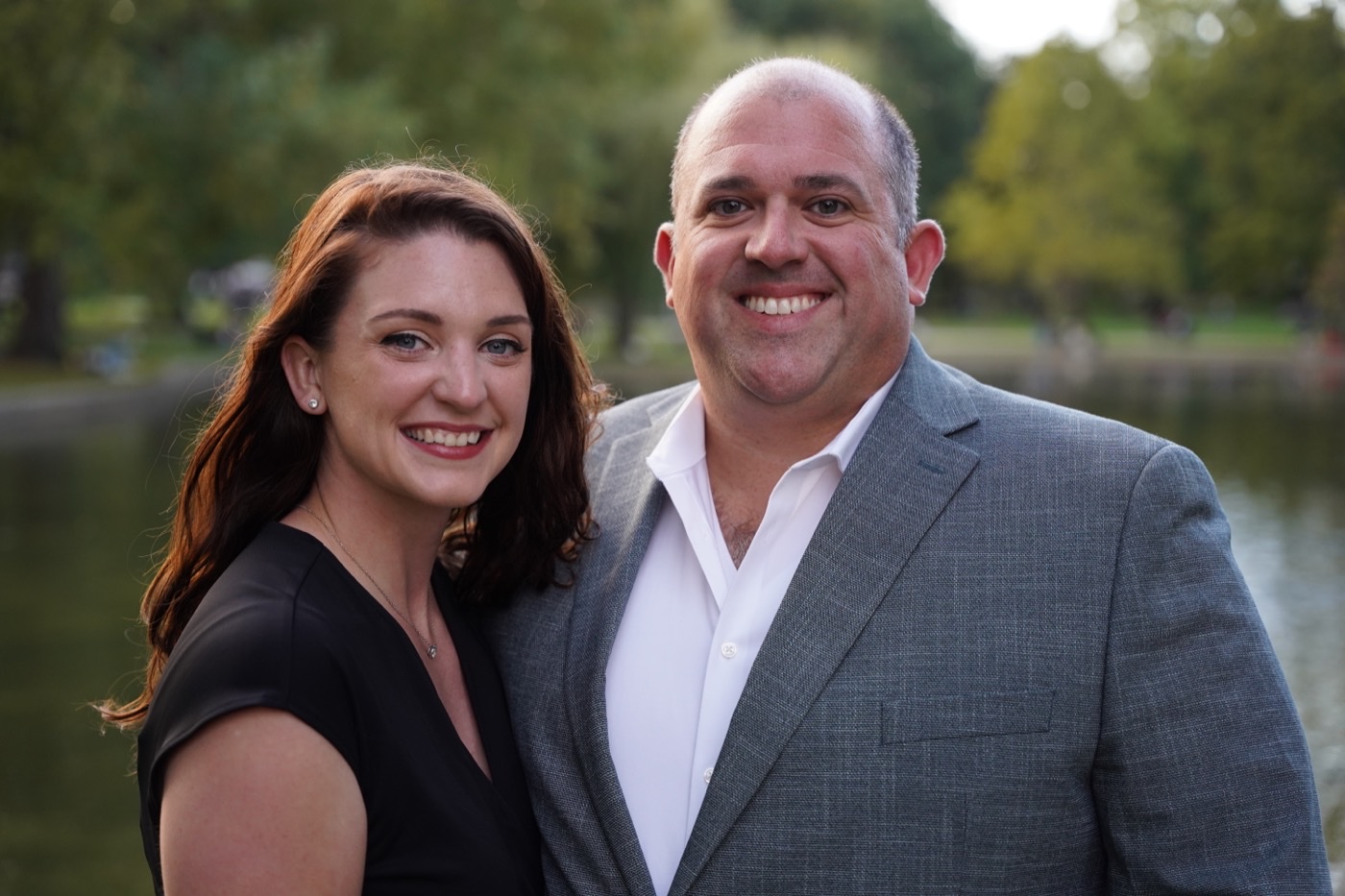 Woman with long auburn hair and a man in a grey suit jacket standing close and smiling at the camera, trees and water behind them.