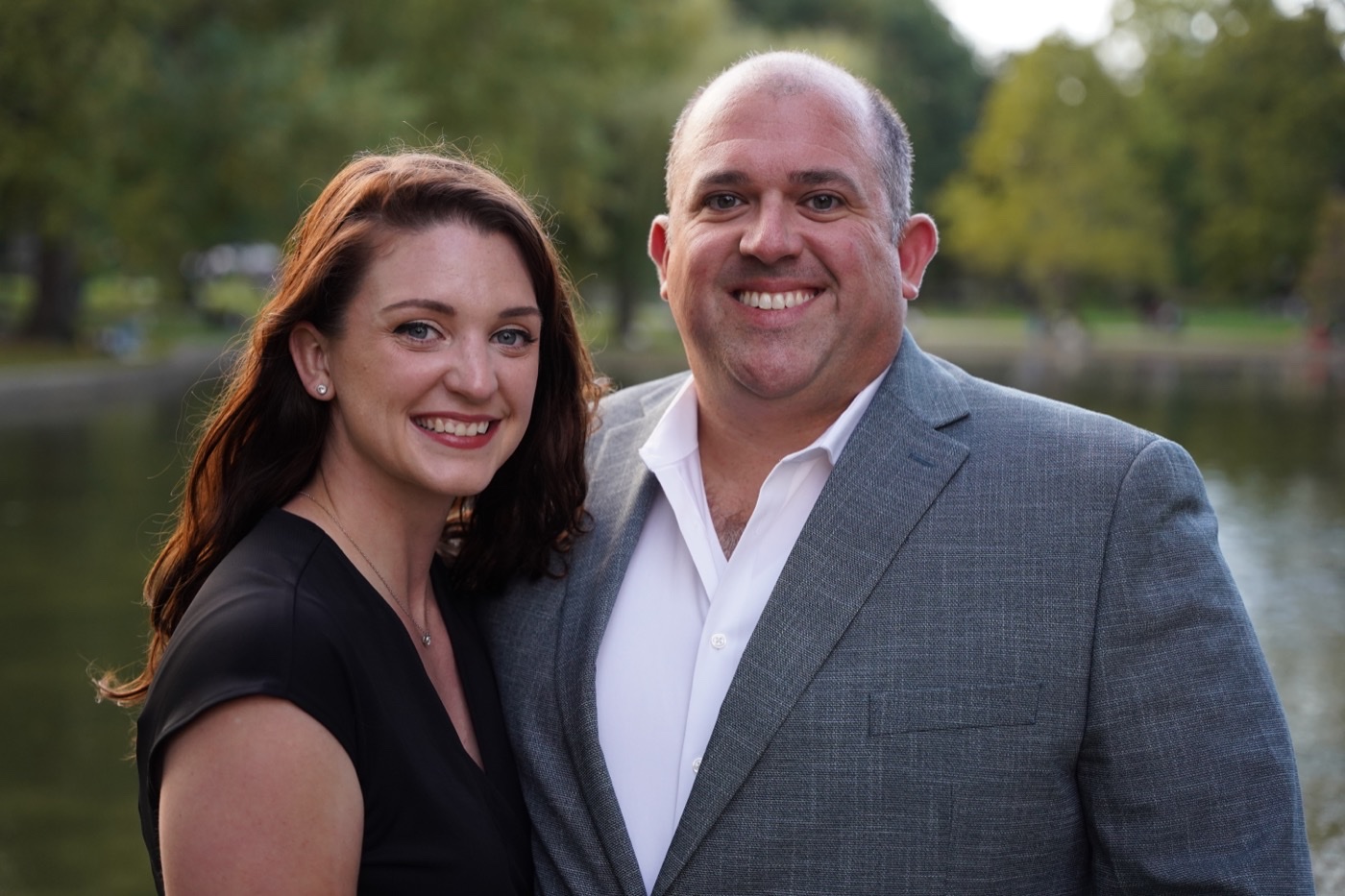 Woman in a black top and a man in a grey suit jacket and white shirt smiling at the camera beside a pond.