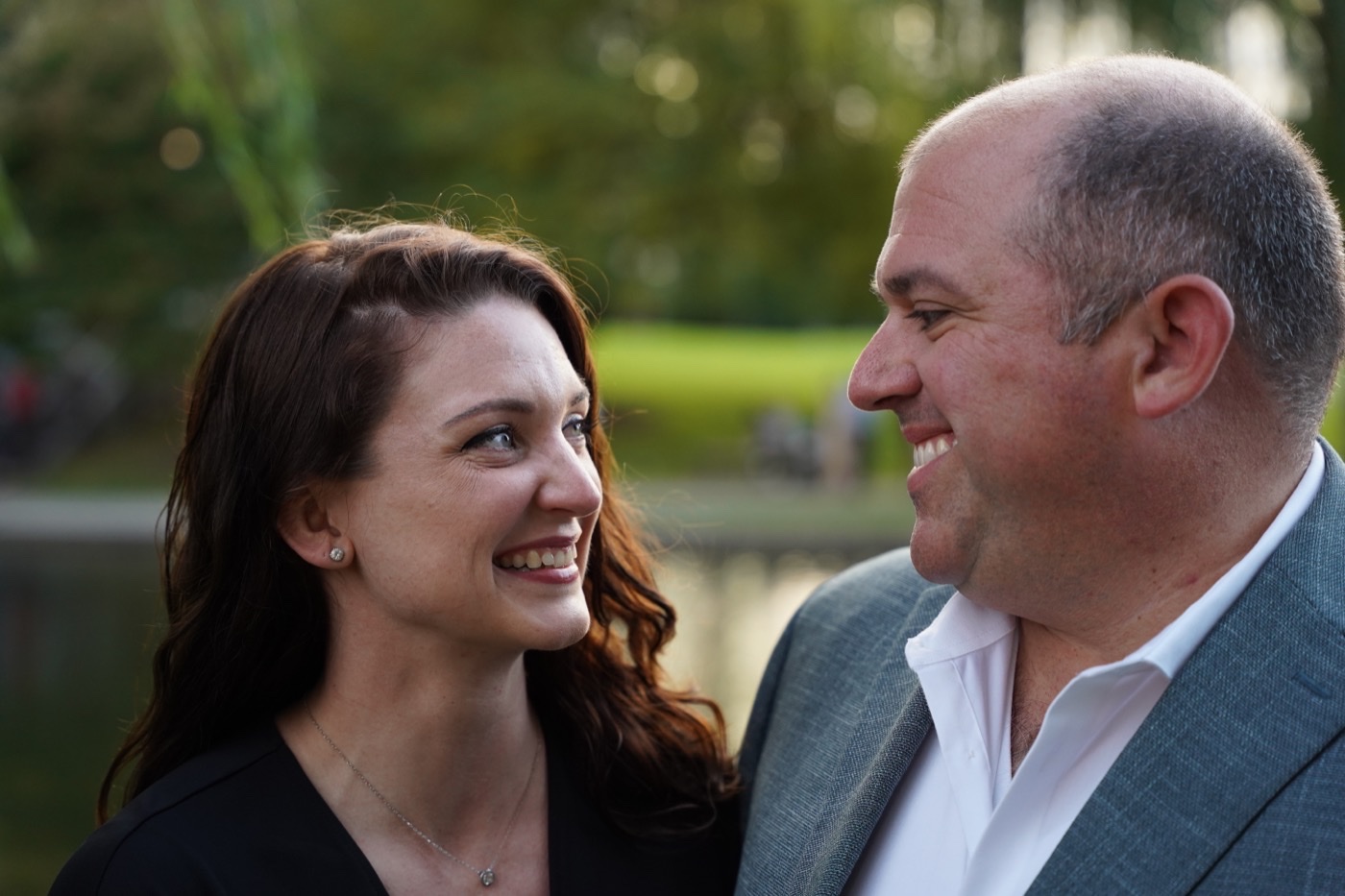 Woman with long auburn hair and a man in a grey suit jacket sharing a laugh, with trees and a pond behind them.