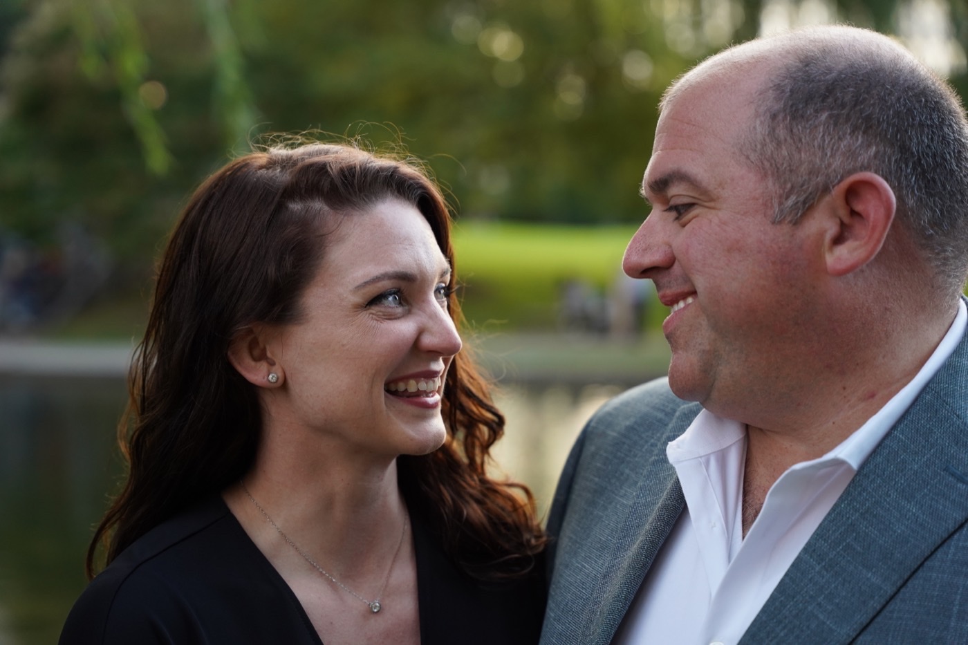 Smiling woman with long auburn hair and a man in a grey suit jacket looking at each other, trees and water behind them.