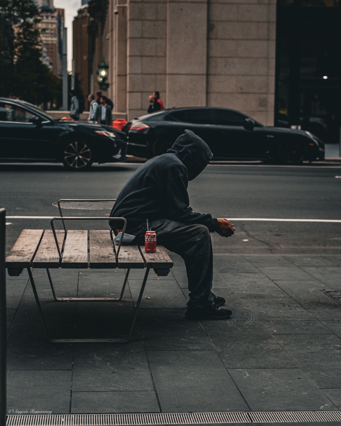 Person in a dark hooded sweatshirt sitting hunched on a wooden bench on a city sidewalk, a soda can beside them.