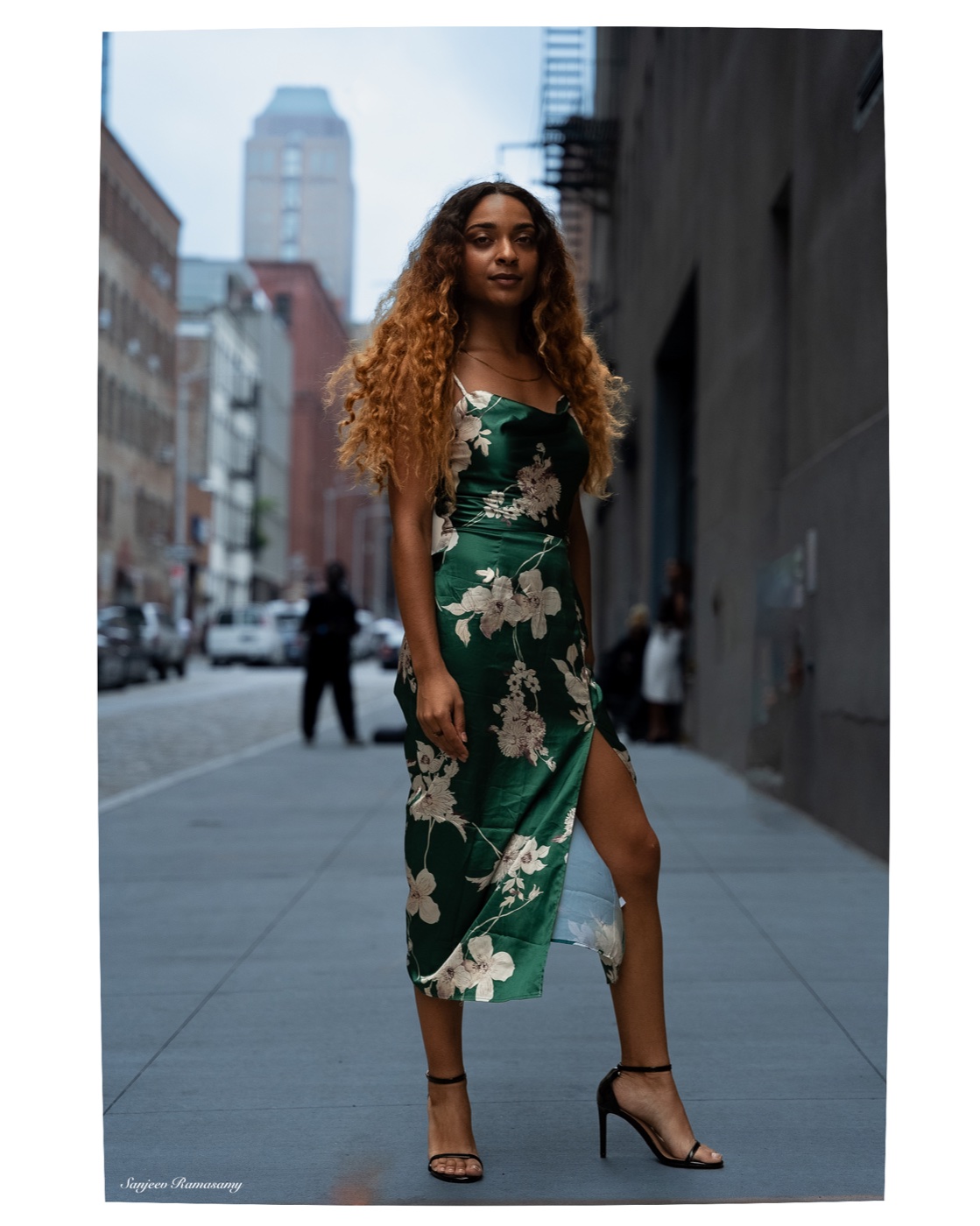 Woman in a green floral slip dress and strappy heels standing on a city sidewalk, a bridge and tall buildings behind her.