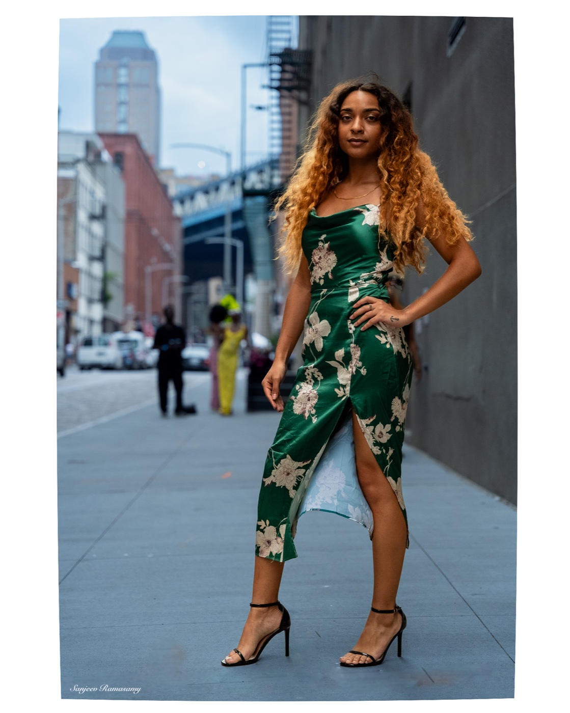 Woman with long wavy hair in a green floral slip dress posing with a hand on her hip on a city sidewalk.