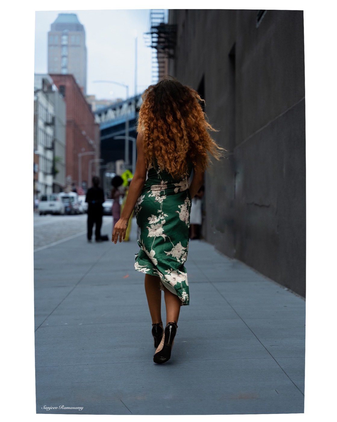 Woman with long wavy auburn hair in a green floral dress walking away down a city sidewalk.