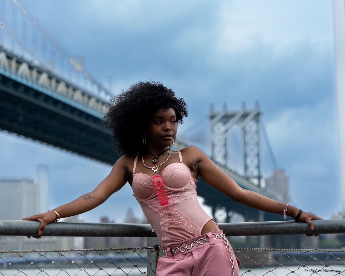 Woman in a pink corset leaning on a railing with her arms stretched wide beneath a large steel bridge and a cloudy sky.