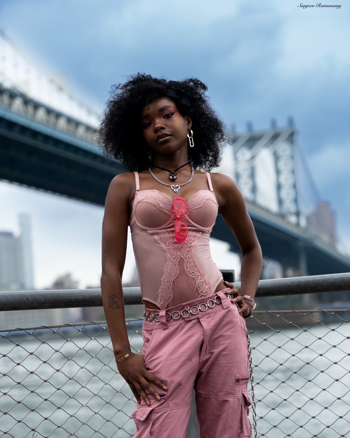 Woman with voluminous curly hair in a pink corset top standing by a railing with one hand on her hip, a large bridge behind her.