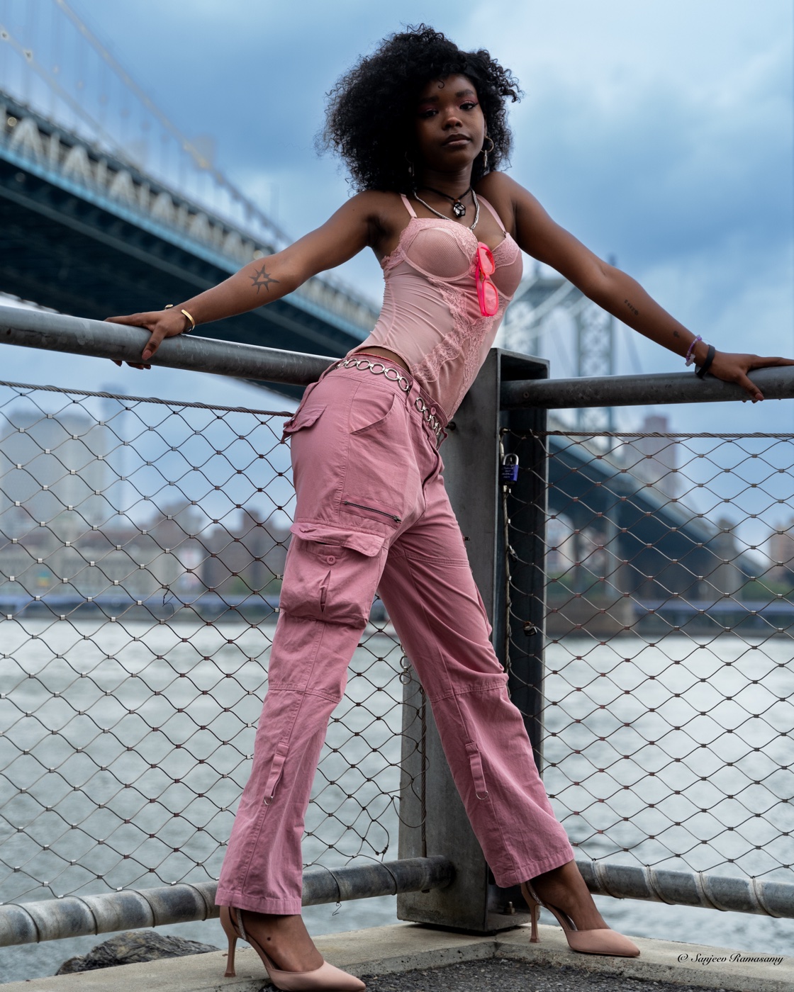 Woman with voluminous curly hair in a pink corset and cargo trousers leaning against a waterfront railing, a steel bridge overhead.