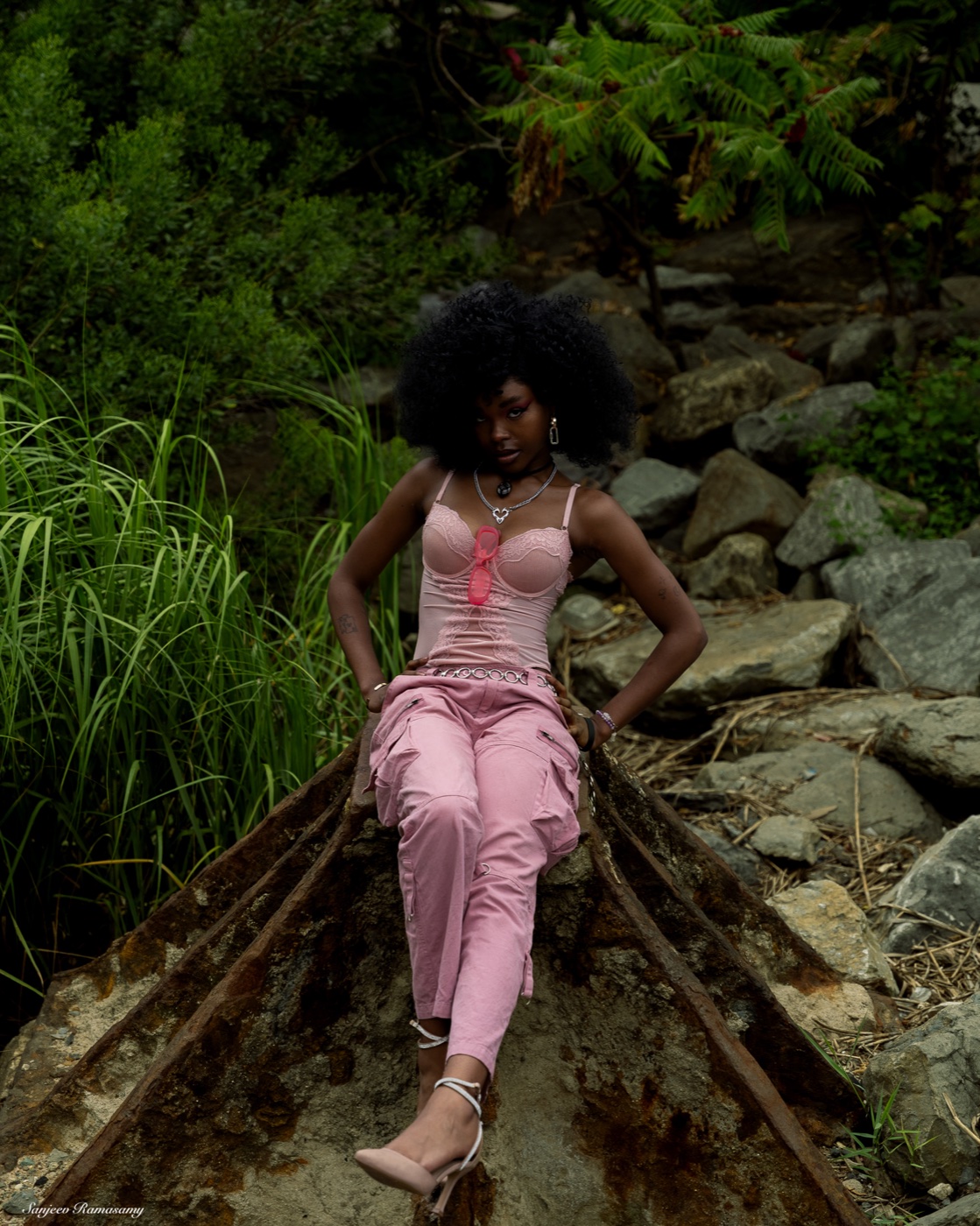 Woman in a pink corset top and pink trousers standing with hands on hips on overgrown rail tracks among rocks and plants.