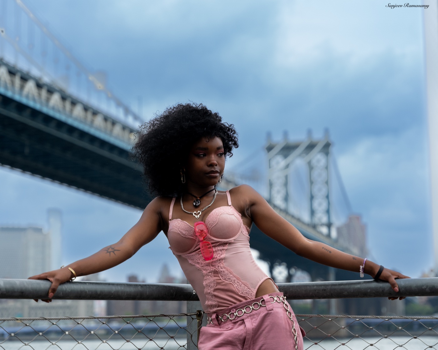 Woman with voluminous curly hair in a pink corset and trousers leaning on a railing beneath a large steel bridge.
