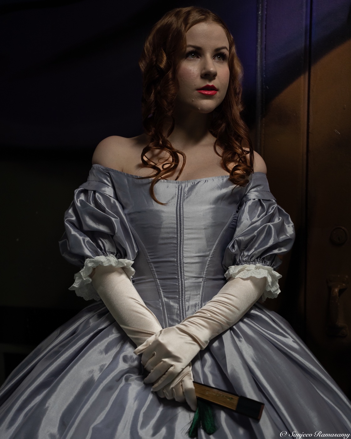 Woman in an off-shoulder silver ball gown and long white gloves with her hands clasped, lit against a dark wall.