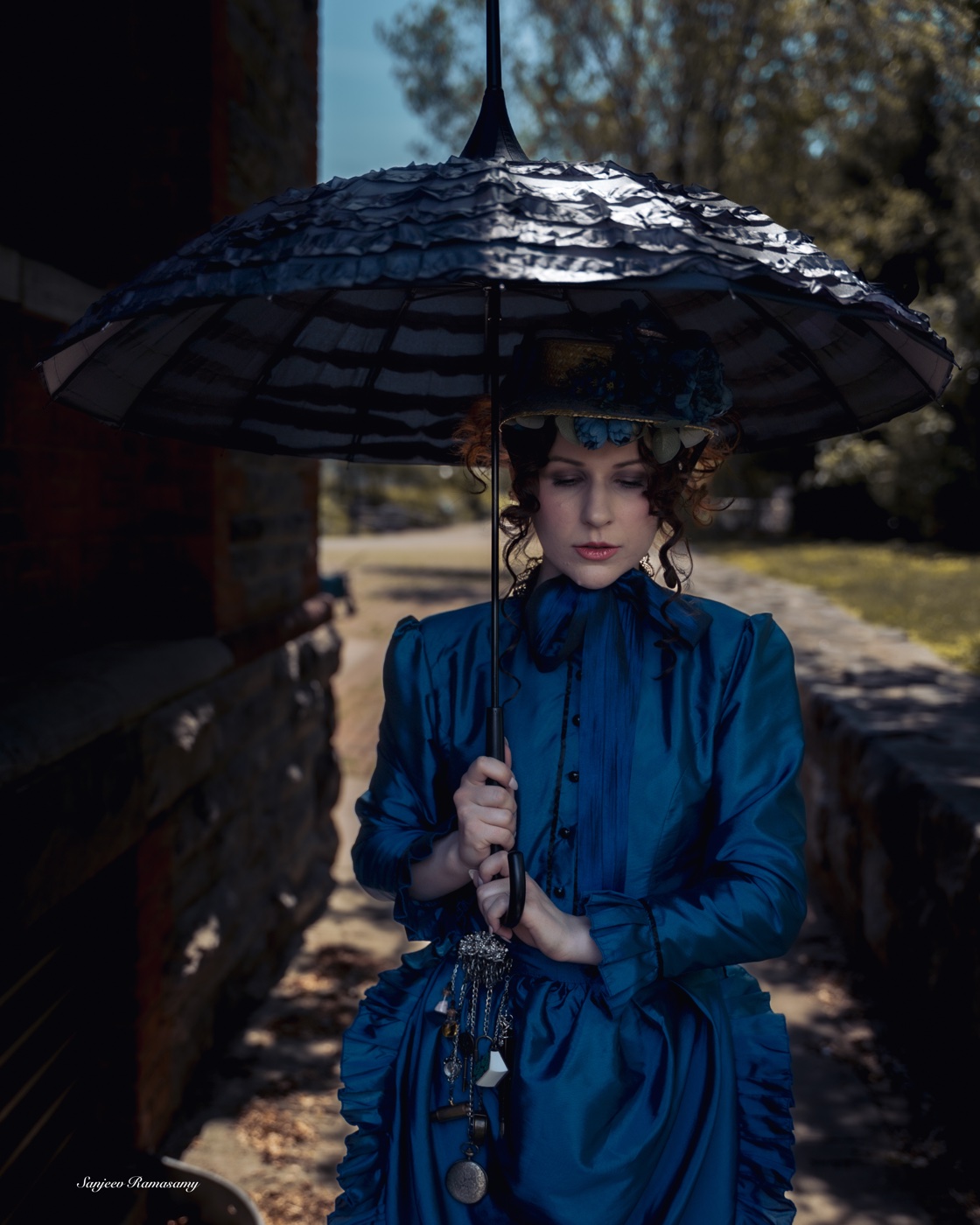 Woman in a blue Victorian gown holding a black ruffled parasol, standing on a garden path between stone walls.