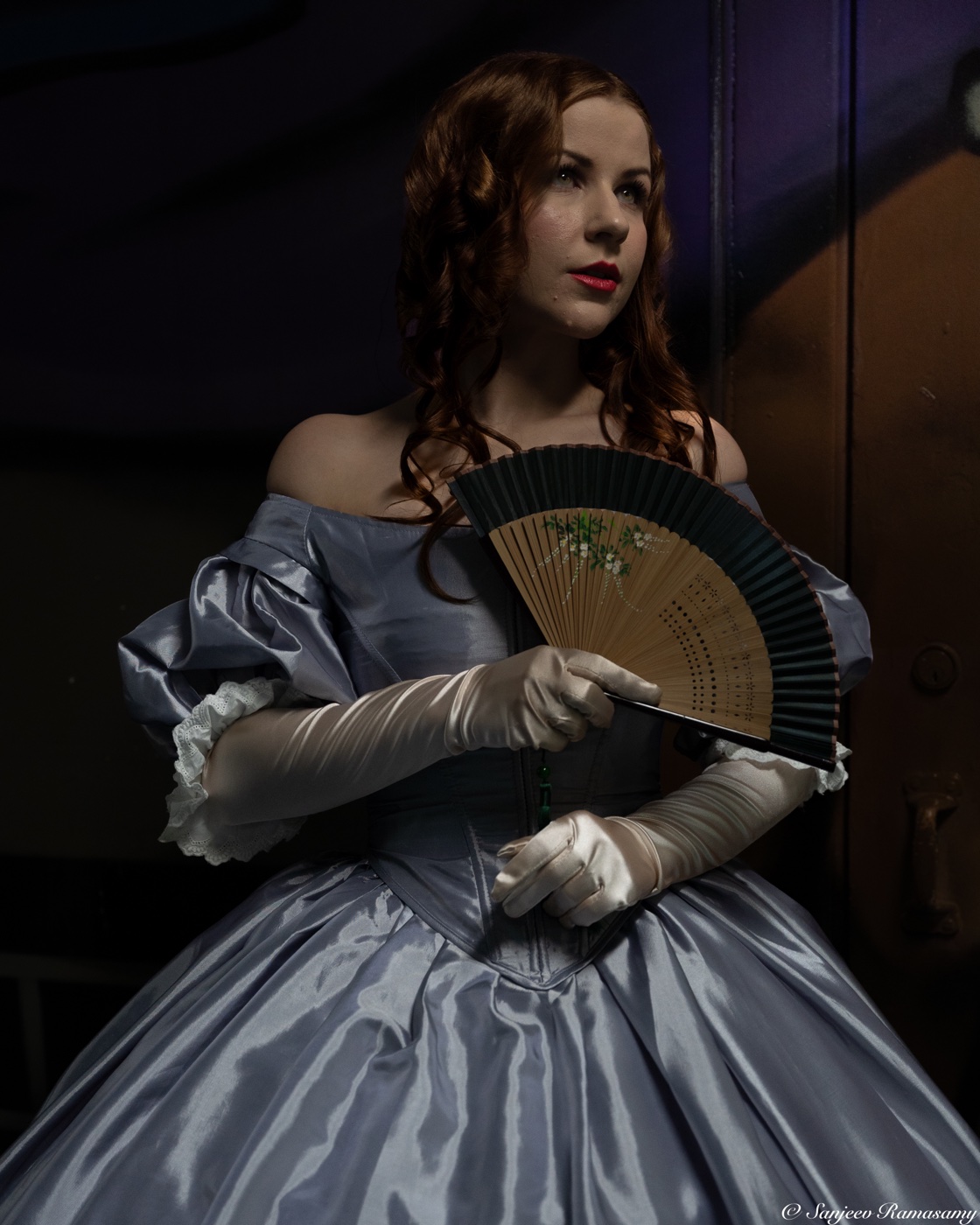 Woman with auburn curls in an off-shoulder silver ball gown and long white gloves, holding a folding fan.