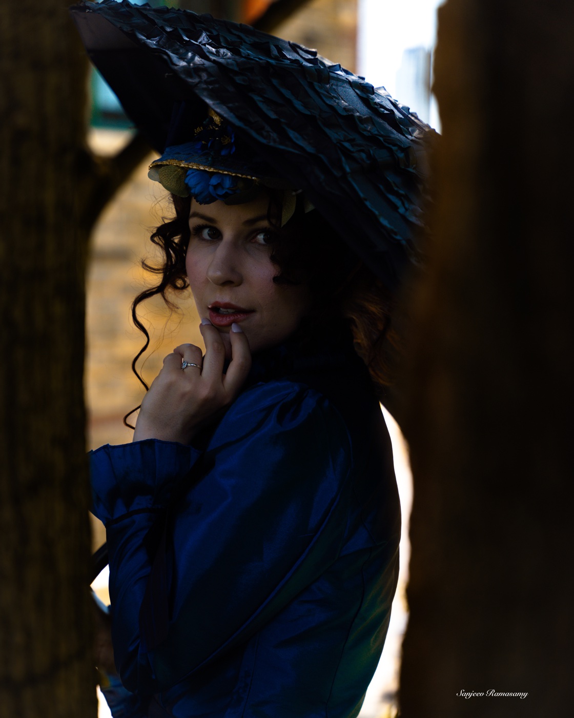 Woman in a blue period gown and hat under a black parasol, half in shadow in a dark doorway.