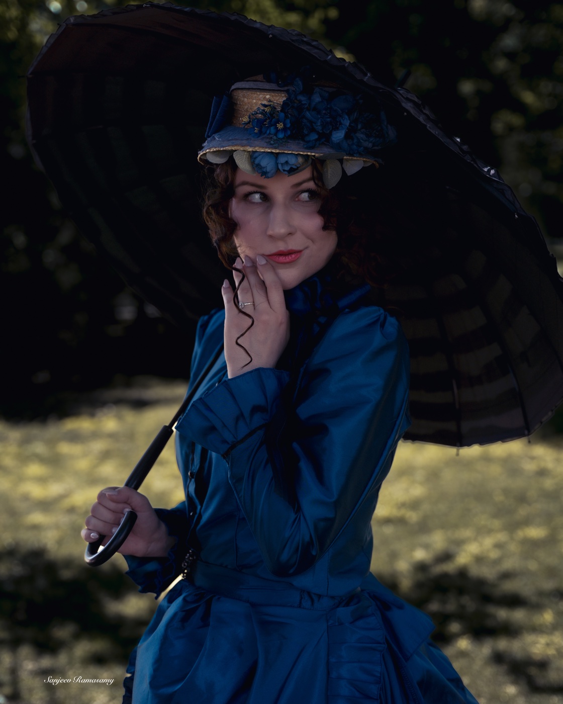 Woman in a blue Victorian gown and feathered hat holding a black parasol over her shoulder, touching her chin.