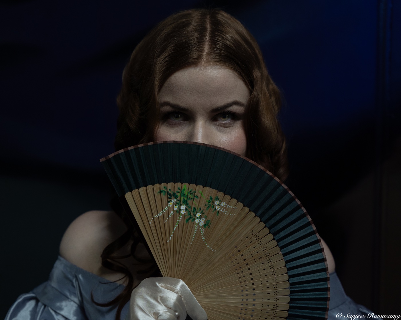 Woman with auburn hair hiding the lower half of her face behind an open painted fan, looking over it at the camera.