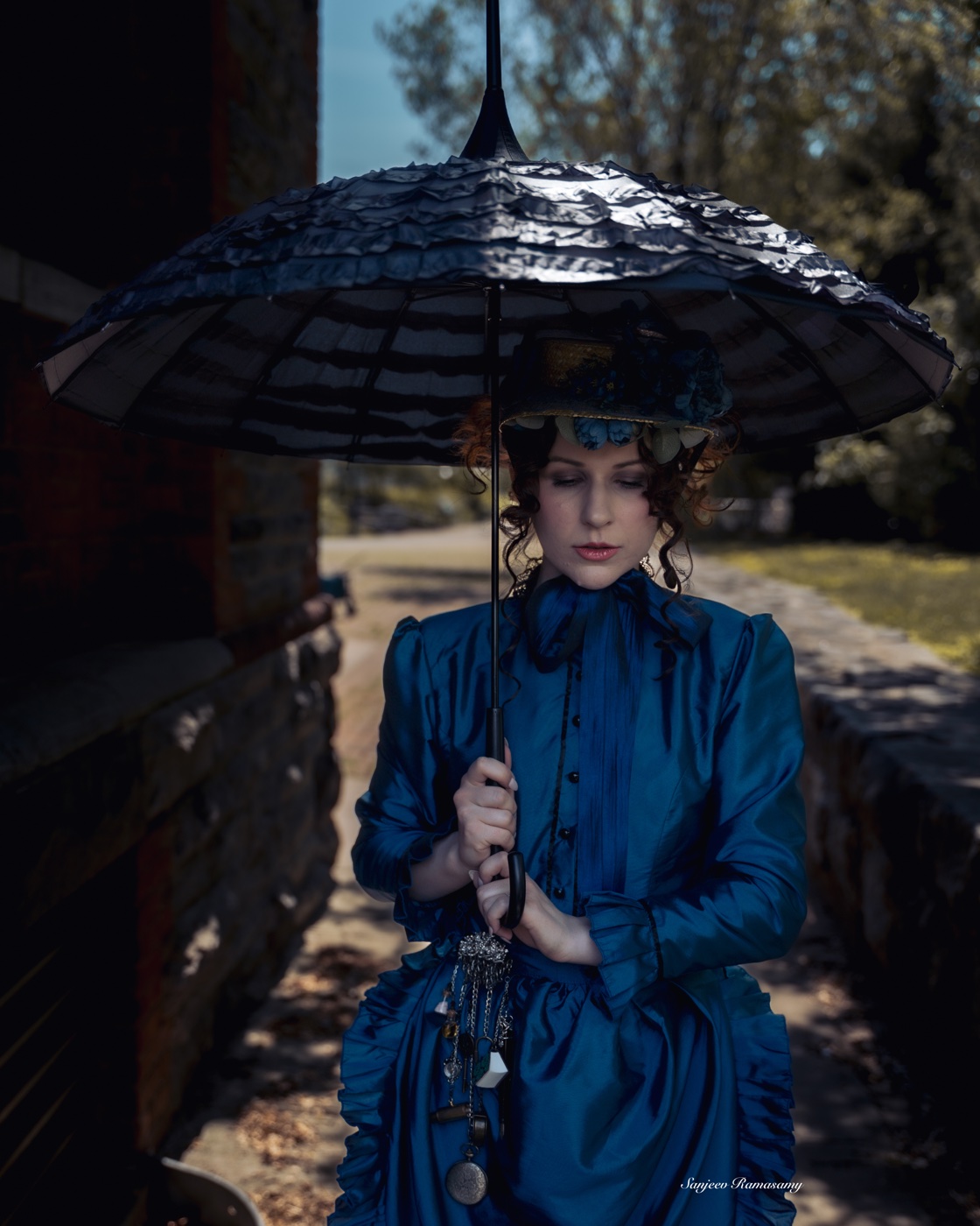 Woman in a Victorian-style blue gown and small hat holding a black ruffled parasol on a shaded garden path.