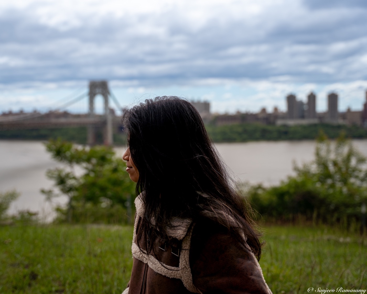 Woman with long dark hair gazing out over a river toward a suspension bridge and a distant skyline under a cloudy sky.
