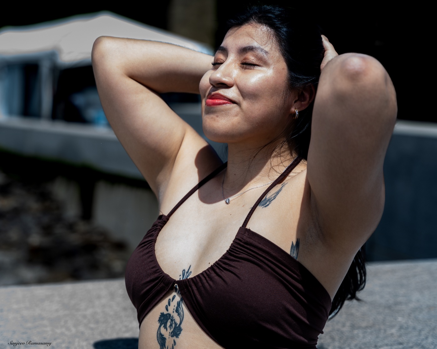 Smiling woman with dark hair and a tattoo, eyes closed and hands behind her head, in bright sunlight.