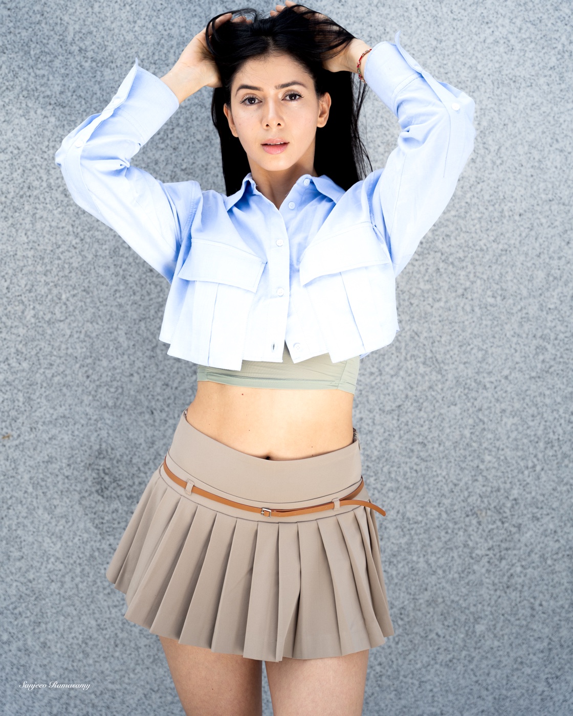Woman in a cropped light-blue shirt and a beige pleated mini skirt posing with arms raised in front of a textured grey wall.