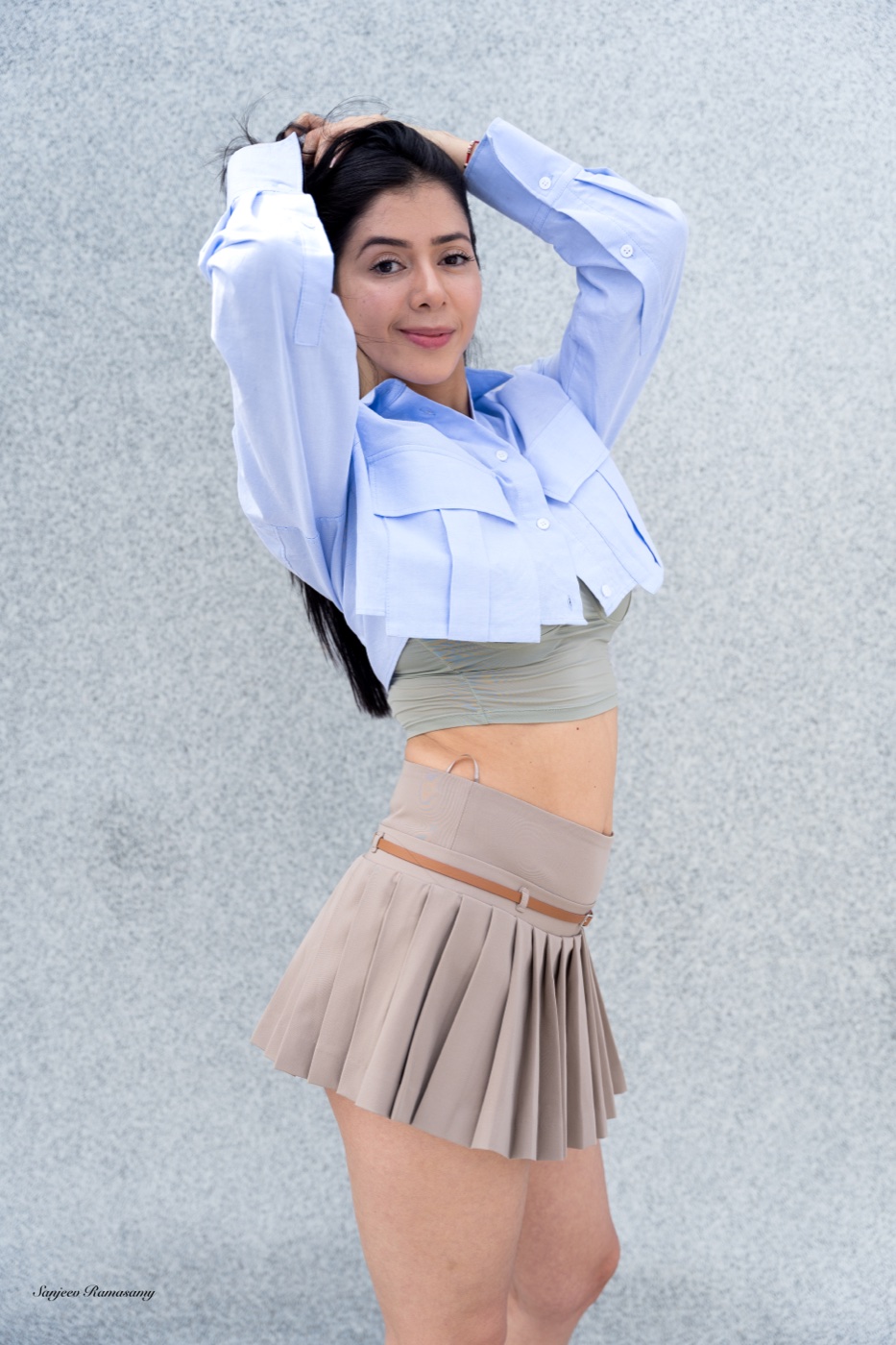 Woman in a cropped light-blue shirt and a beige pleated skirt posing with her hands behind her head against a grey wall.